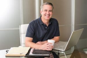 Smiling entrepreneur holding a coffee seated at a desk with laptop and open ledgers; tax planning.