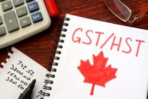 GST/HST written in a notebook on a wooden desk with pen, notes, and calculator for business considering requirements to register for GST and register for HST