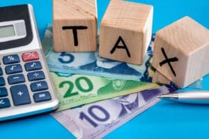 Wooden cubes that spell out the word “tax” sit on top of a trio of Canadian bills next to a calculator and a pen.